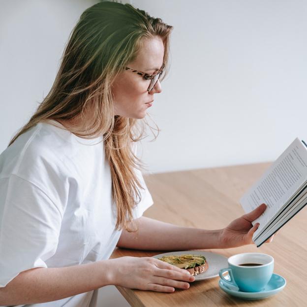 Mujer leyendo un libro mientras toma una tostada de aguacate y un café. 